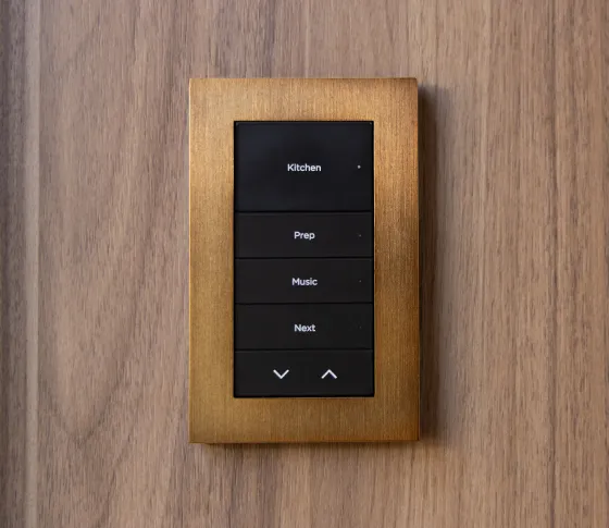 Lux keypad with dark buttons and a warm brass-tone metal faceplate mounted on wood; buttons engraved Kitchen, Prep, Music, Next, Down / Up arrow icons